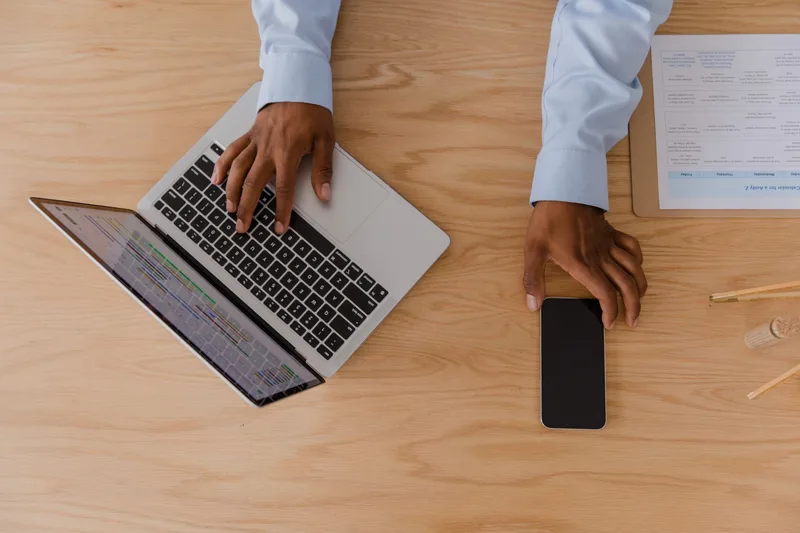 Tangan menggunakan laptop menunjukkan jadual berwarna di skrin, dengan telefon pintar di sebelah untuk semakan jadual kerja