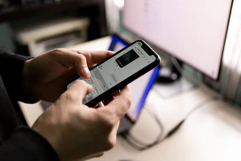 Person typing a message on a smartphone at a desk with a computer monitor behind it