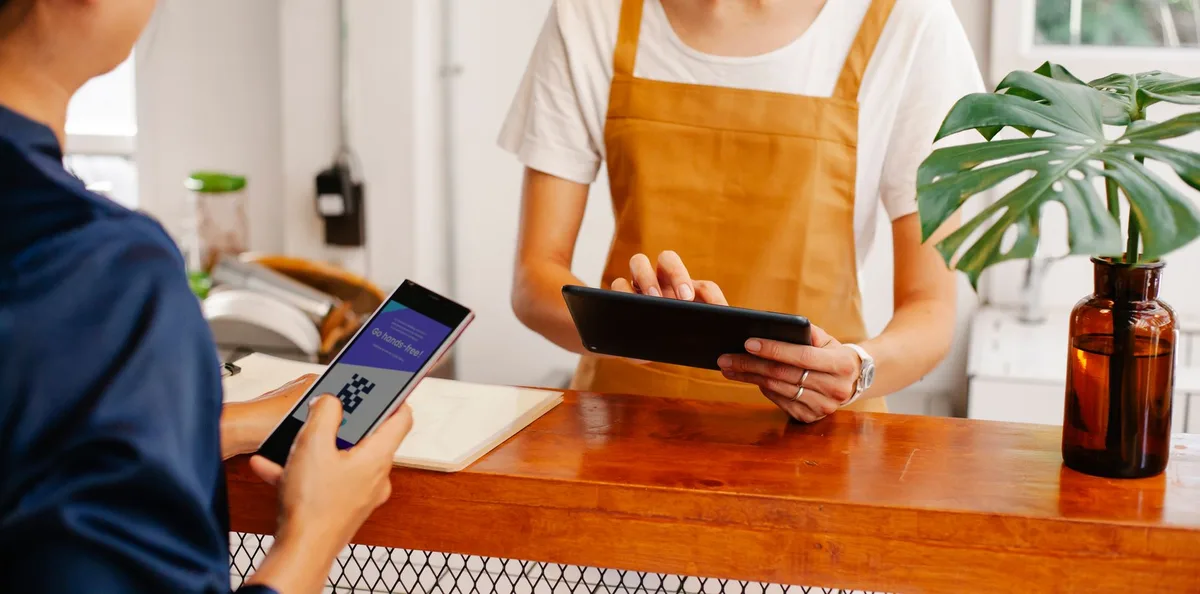 Customer holding a phone with a QR code while a staff member in an apron uses a tablet at a shop counter