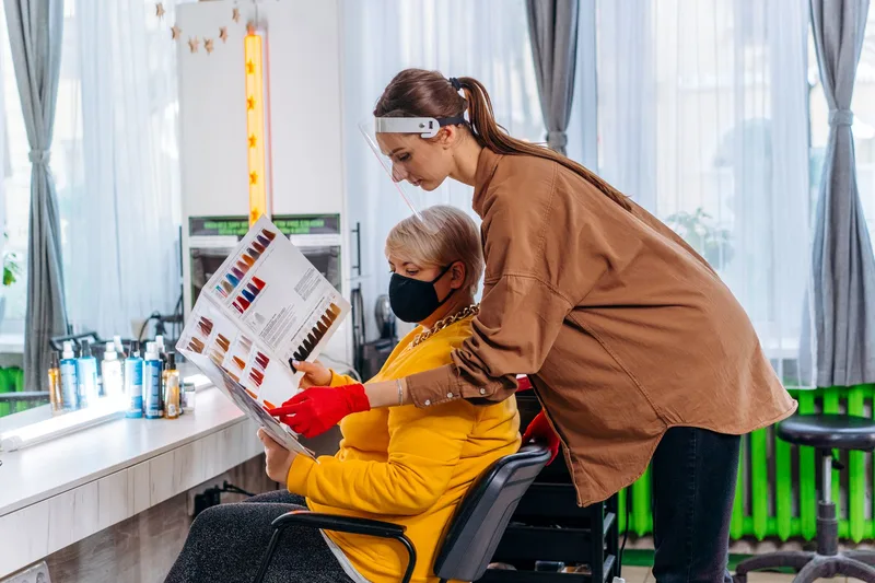 Hairdresser in a face shield showing a hair colour chart to a client in a salon chair